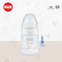 Le FCP1-Biberon NUK en plastique 150ml présente un bouchon et une tétine transparents, des motifs en forme de cœur, les logos NUK Air System et BPA-free, ainsi qu'une icône de contrôle de la température sur un fond beige et blanc avec des illustrations en forme de cœur.